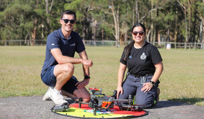 Two smiling trainees kneel beside a quadcopter drone on a landing pad in an open field during UAV training.