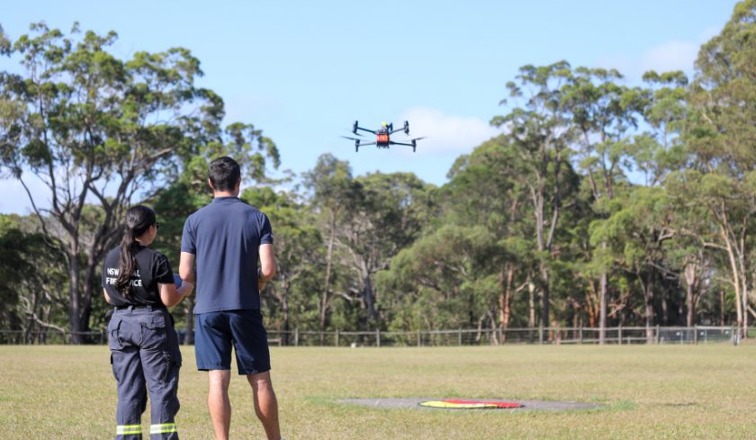 Two people stand in a grassy field operating a quadcopter drone during training, with trees in the background.