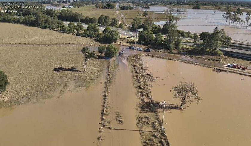 Drone view of floodwaters covering a rural road, with vehicles stopped at a submerged crossing and fields inundated.