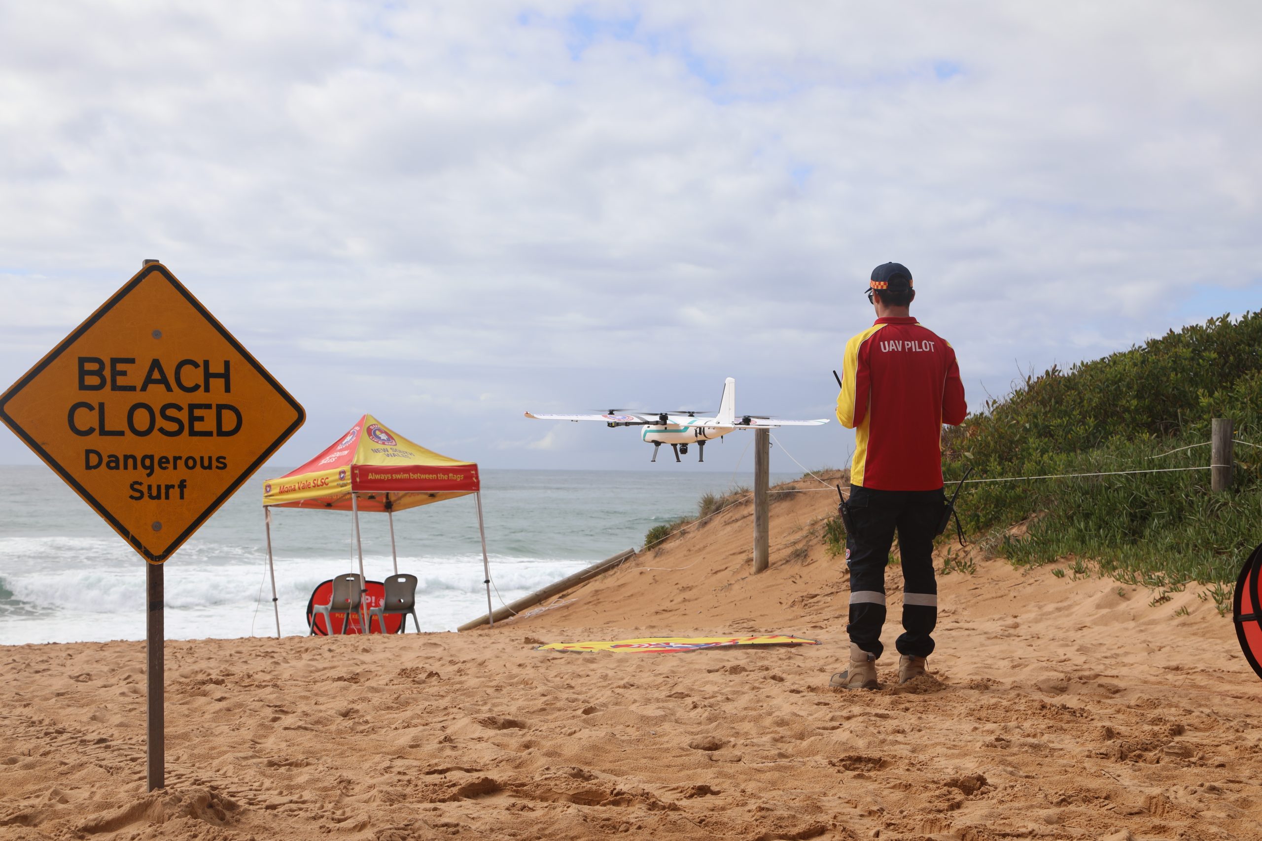 A sandy beach scene with a yellow warning sign in the foreground reading “BEACH CLOSED – Dangerous Surf.” Behind the sign, a red-and-yellow Surf Life Saving NSW tent is set up near the shoreline with two empty chairs underneath. To the right, a person wearing a red and yellow shirt labeled “UAV PILOT” on the back stands on the sand, operating a remote controller and facing the ocean. A white drone with multiple propellers hovers in mid-air in front of the person. The background shows choppy waves, a cloudy sky, and low green vegetation along the dune.